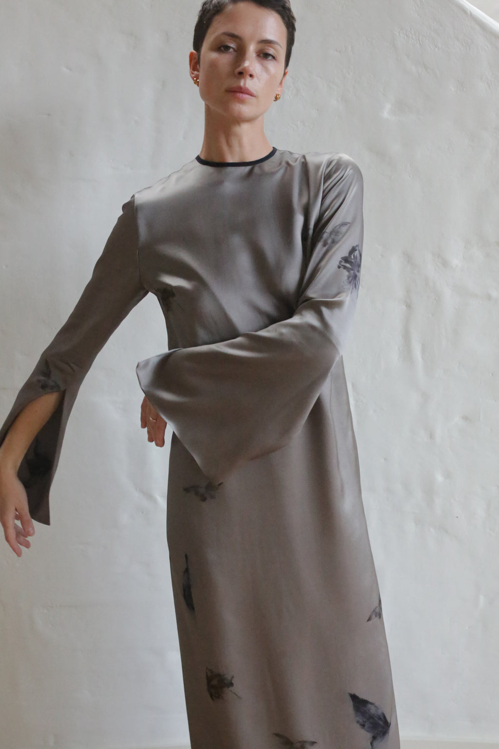 A person with short hair wears The Butterfly Dress | LJ X CL Printed Silk—a long, silvery-gray silk dress with flared sleeves and a subtle dark butterfly print—standing against a plain, light-colored textured wall.