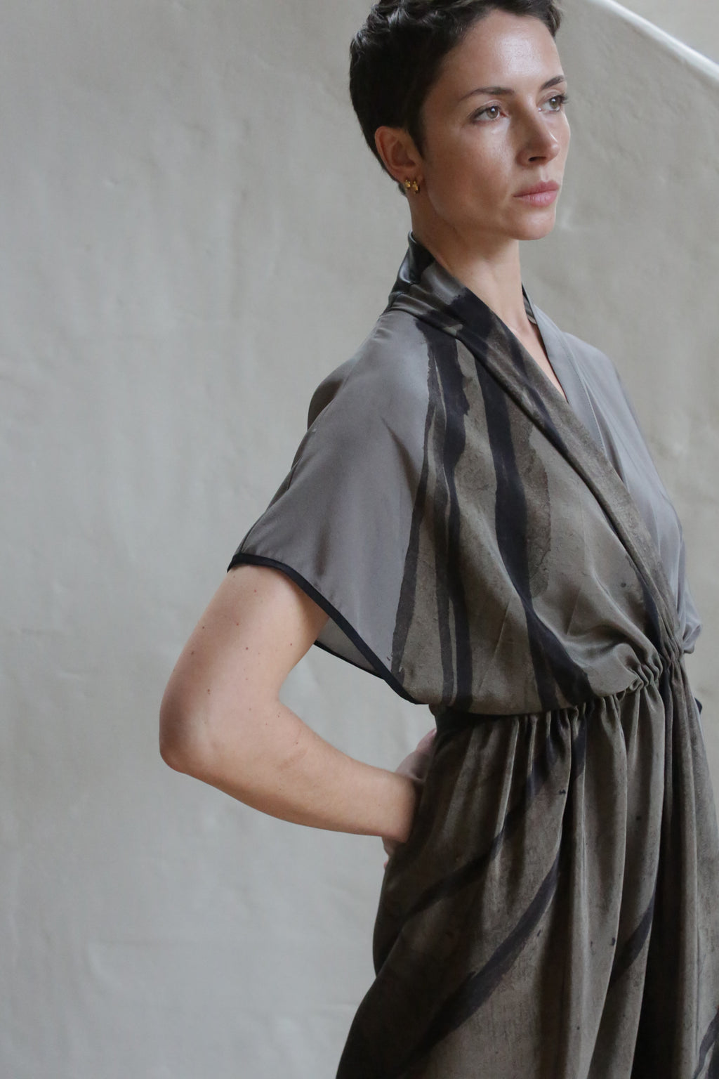 A woman with short dark hair stands against a neutral background, wearing the Lune Dress | Butterfly Wing—a loose-fitting, short-sleeved dress with abstract dark patterns. She gazes thoughtfully into the distance, one hand resting on her hip.