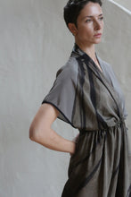 Load image into Gallery viewer, A woman with short dark hair stands against a neutral background, wearing the Lune Dress | Butterfly Wing—a loose-fitting, short-sleeved dress with abstract dark patterns. She gazes thoughtfully into the distance, one hand resting on her hip.