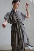 A woman with short dark hair wears the Lune Dress | Butterfly Wing, a loose gray kimono-style dress with black abstract patterns. She stands indoors against a light wall, one arm raised and eyes gently closed.