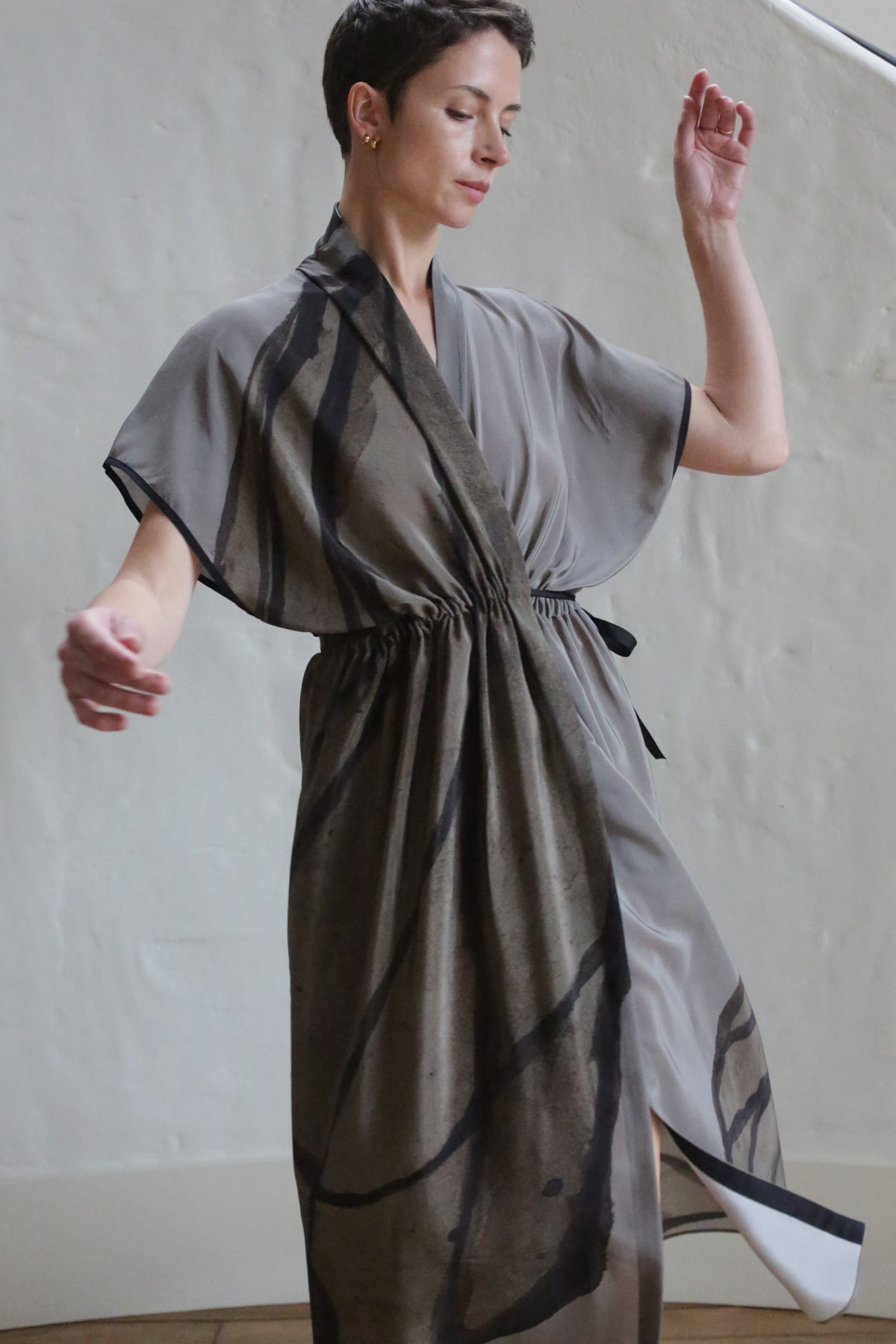 A woman with short dark hair wears the Lune Dress | Butterfly Wing, a loose gray kimono-style dress with black abstract patterns. She stands indoors against a light wall, one arm raised and eyes gently closed.