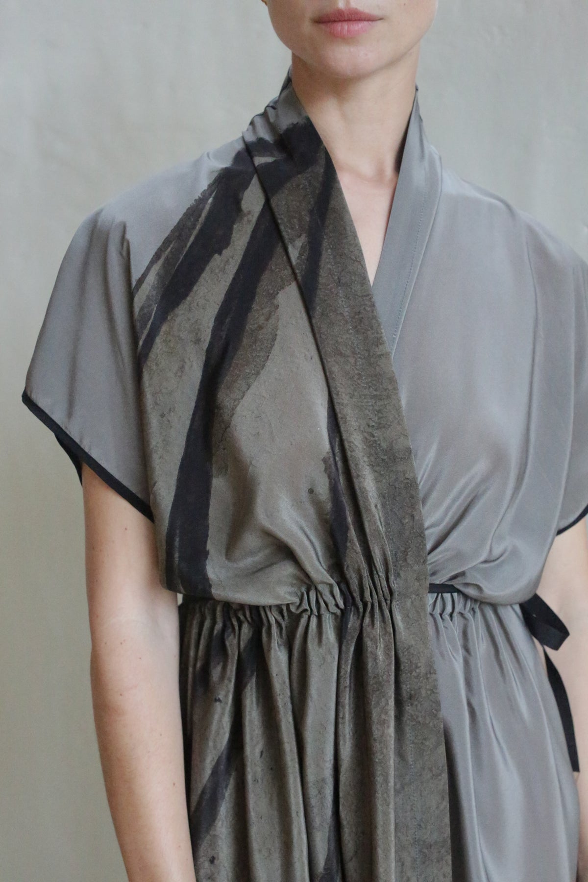 A person is wearing the Lune Dress | Butterfly Wing, a grey short-sleeved dress with a black abstract pattern and gathered waist, standing against a plain light background. Only their upper body and lower face are visible.