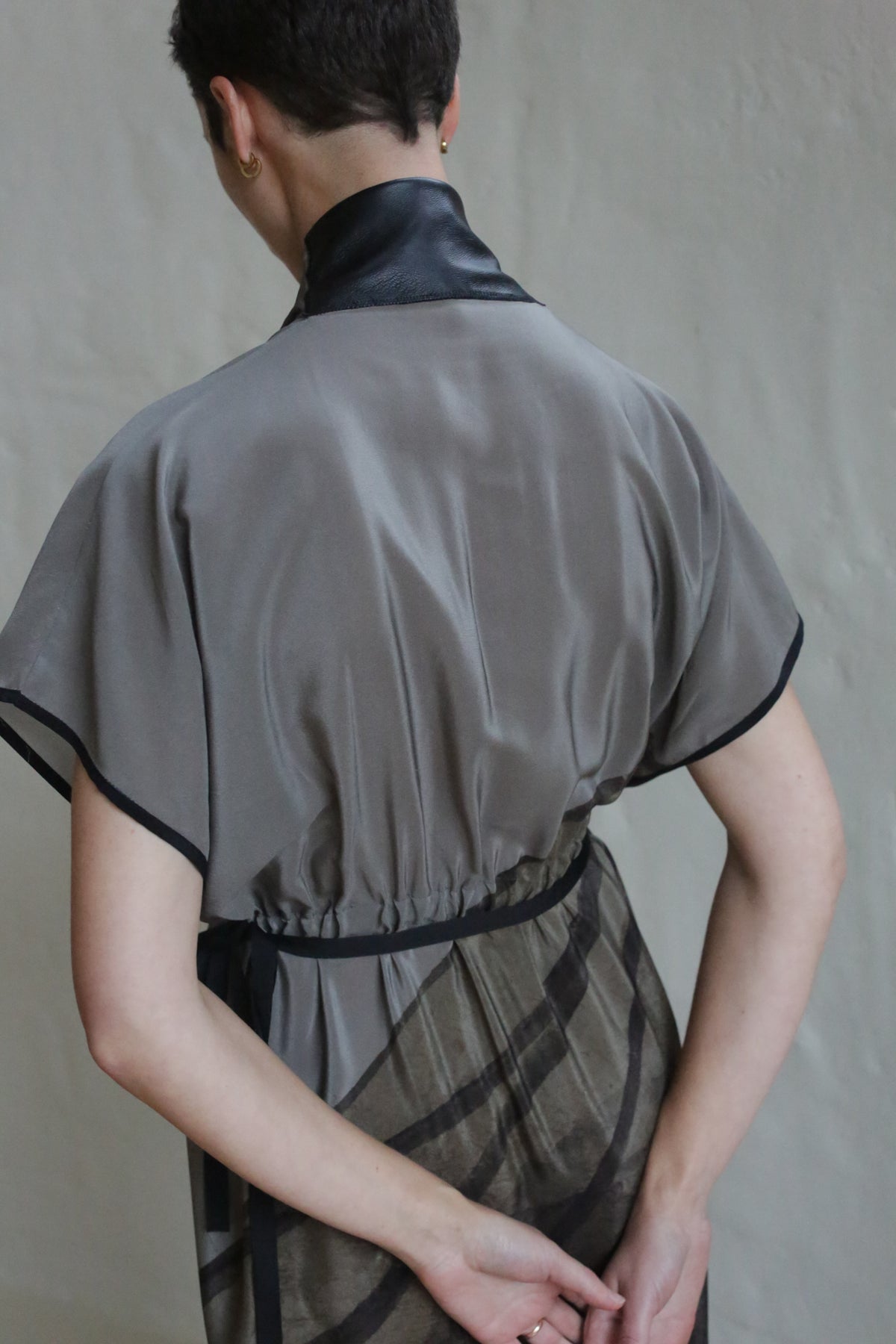 A person with short hair faces away, wearing the Lune Dress | Butterfly Wing—a sheer gray dress with black trim and a shiny black collar. Their arms are bent behind their back, set against a plain light wall.