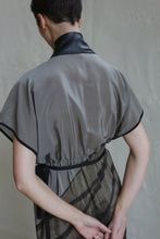 Load image into Gallery viewer, A person with short hair faces away, wearing the Lune Dress | Butterfly Wing—a sheer gray dress with black trim and a shiny black collar. Their arms are bent behind their back, set against a plain light wall.