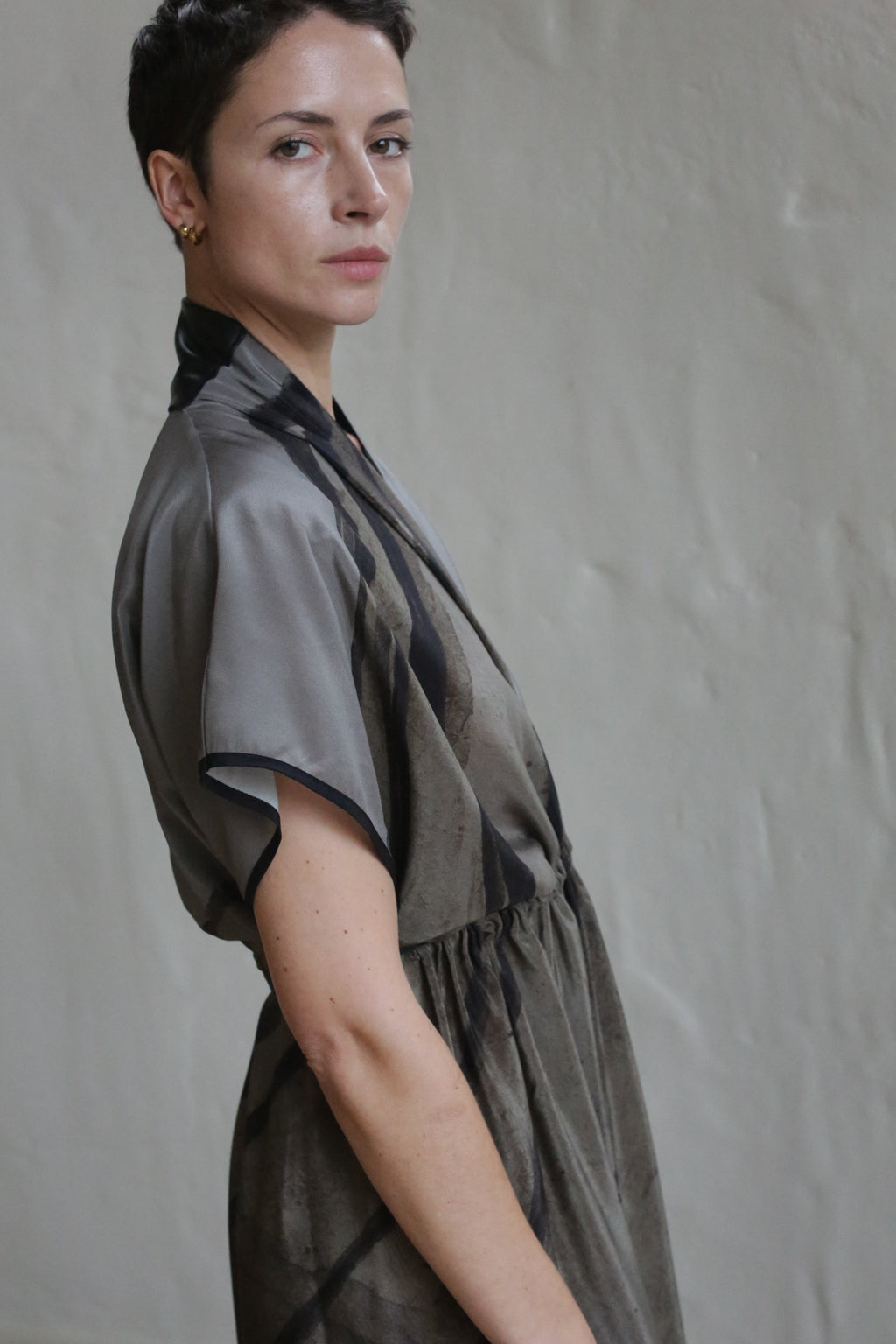 A person with short dark hair and gold earrings wears the Lune Dress | Butterfly Wing, a gray and black short-sleeved dress, standing sideways and looking at the camera against a light-colored wall.
