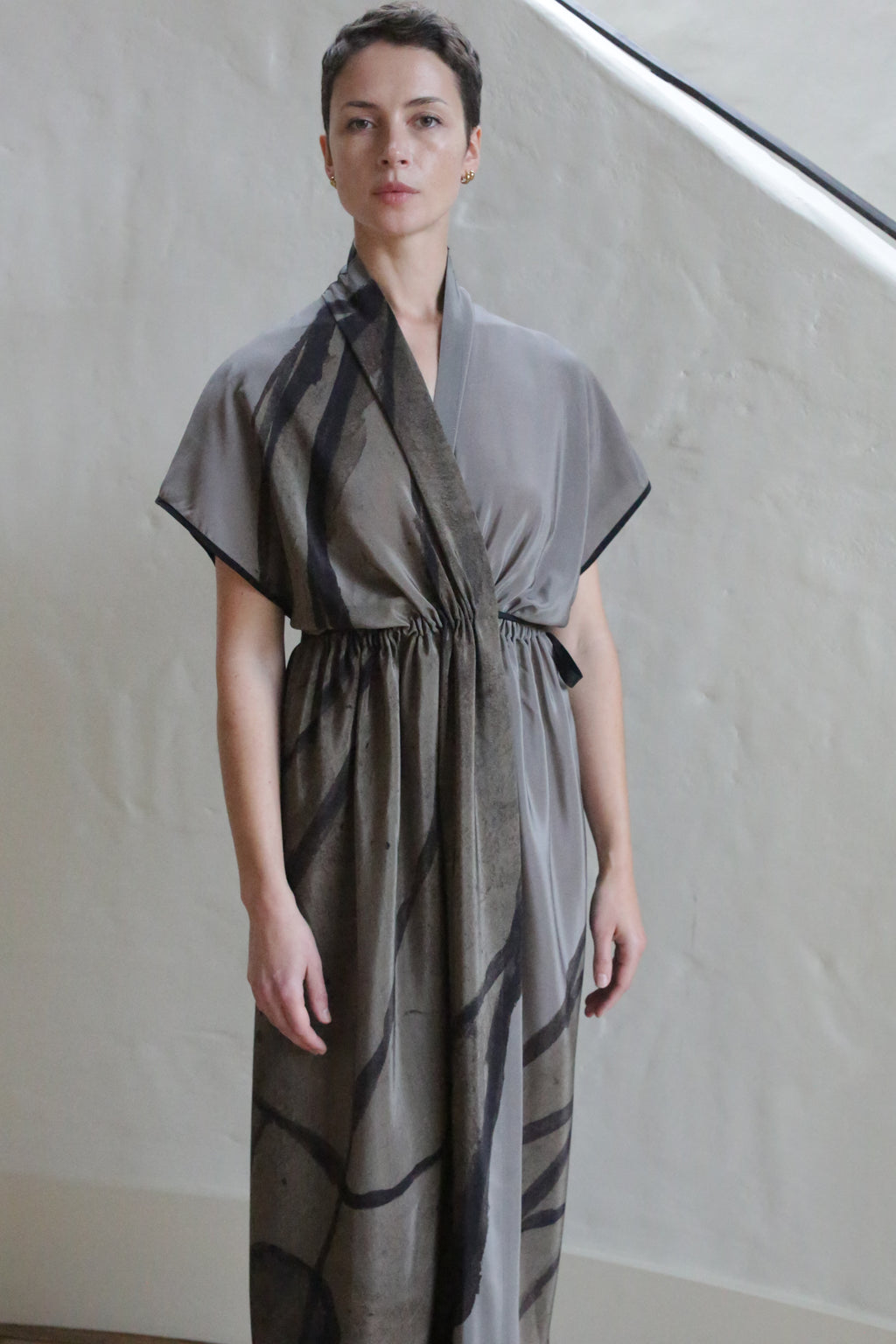 A person with short hair wears the Lune Dress | Butterfly Wing, a loose grey wrap dress with black abstract lines, standing indoors by a light-colored, textured wall and staircase.