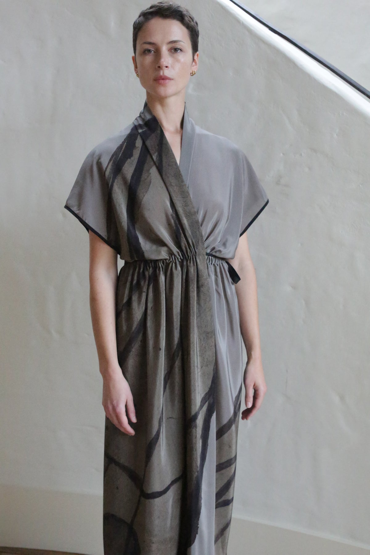 A person with short hair wears the Lune Dress | Butterfly Wing, a loose grey wrap dress with black abstract lines, standing indoors by a light-colored, textured wall and staircase.