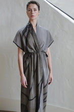 Load image into Gallery viewer, A person with short hair wears the Lune Dress | Butterfly Wing, a loose grey wrap dress with black abstract lines, standing indoors by a light-colored, textured wall and staircase.