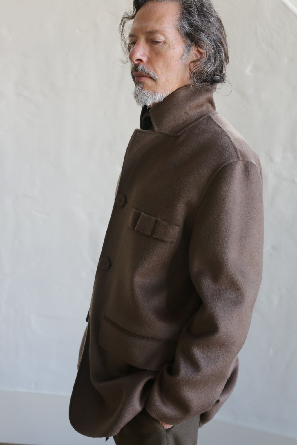 A man with wavy hair and a mustache stands with his eyes closed, hands in pockets, wearing the Sculptor's Jacket | Double Face Cashmere Taupe, in front of a light-colored textured wall.
