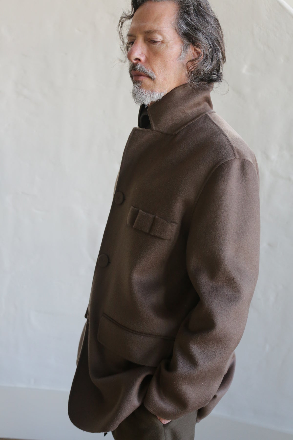 A man with wavy hair and a mustache stands with his eyes closed, hands in pockets, wearing the Sculptor's Jacket | Double Face Cashmere Taupe, in front of a light-colored textured wall.