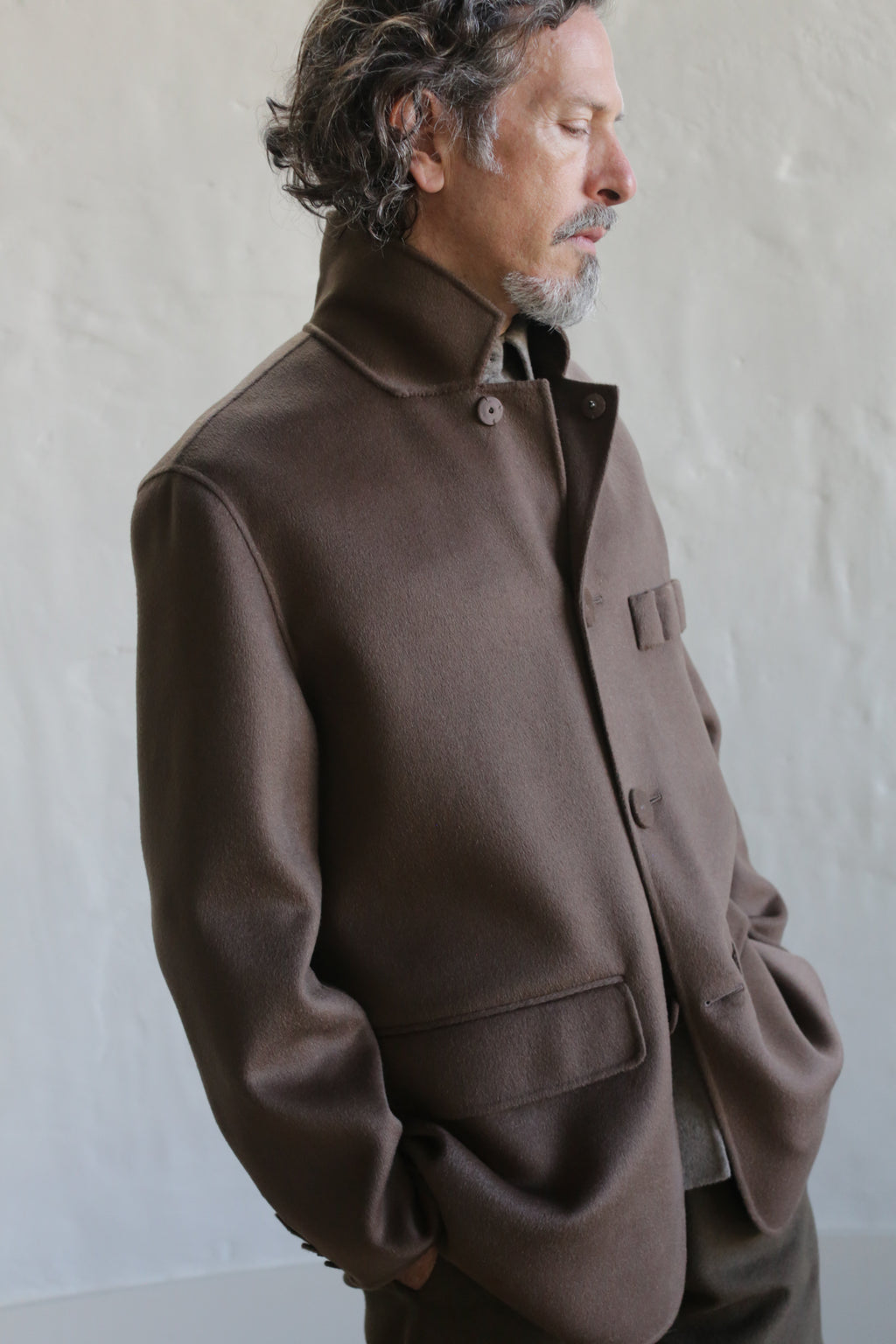 A man with wavy hair and a beard wears the Sculptor's Jacket in Double Face Cashmere Taupe, buttoned up as he stands against a light gray textured wall, looking down thoughtfully with his hands in his pockets.