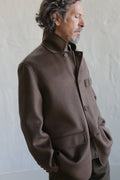 A man with wavy hair and a beard wears the Sculptor's Jacket in Double Face Cashmere Taupe, buttoned up as he stands against a light gray textured wall, looking down thoughtfully with his hands in his pockets.