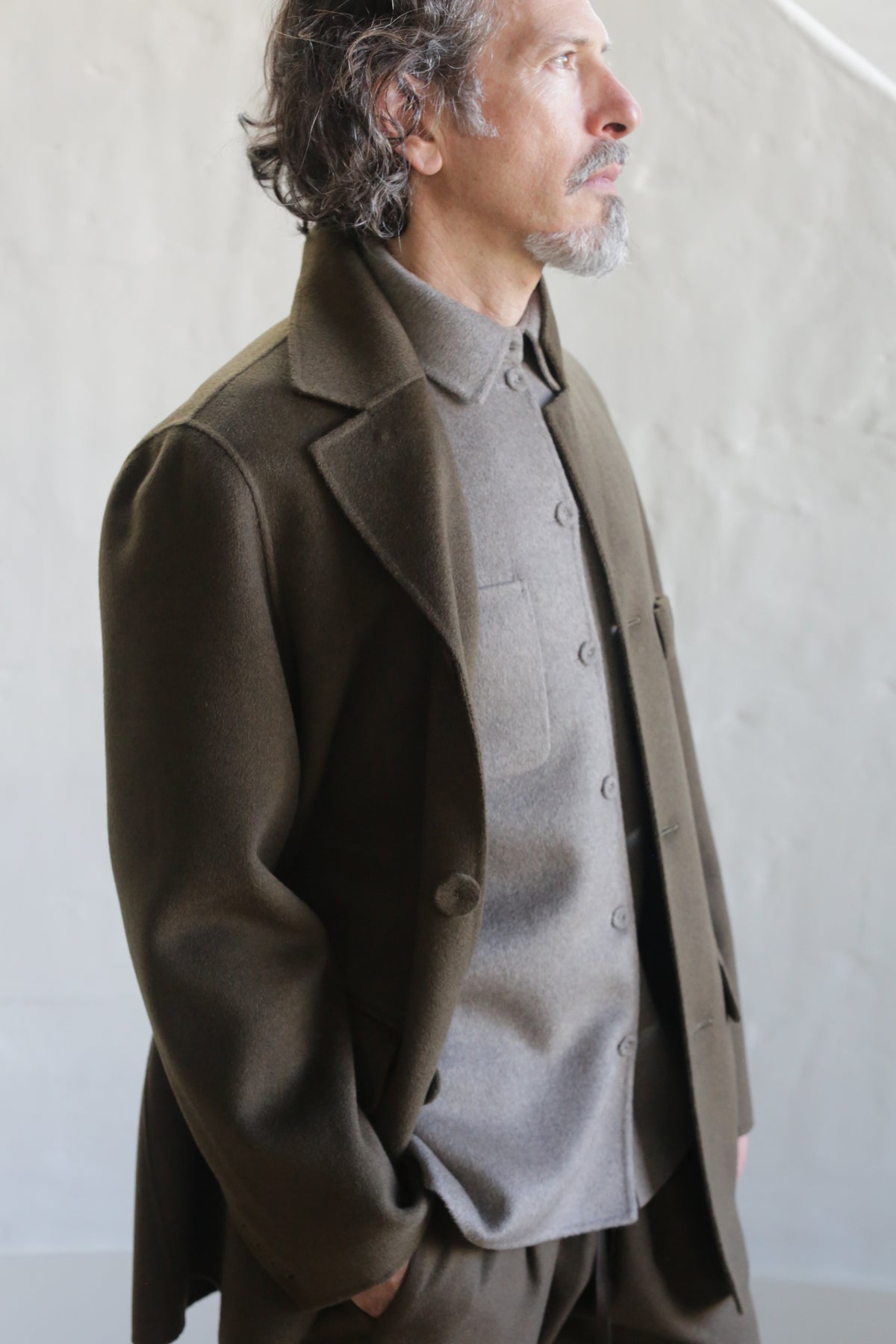 A man with wavy hair and a goatee stands in profile, wearing the Sculptor's Jacket in olive double face cashmere over a gray button-up shirt, his hands in his pockets, set against a light-colored wall.