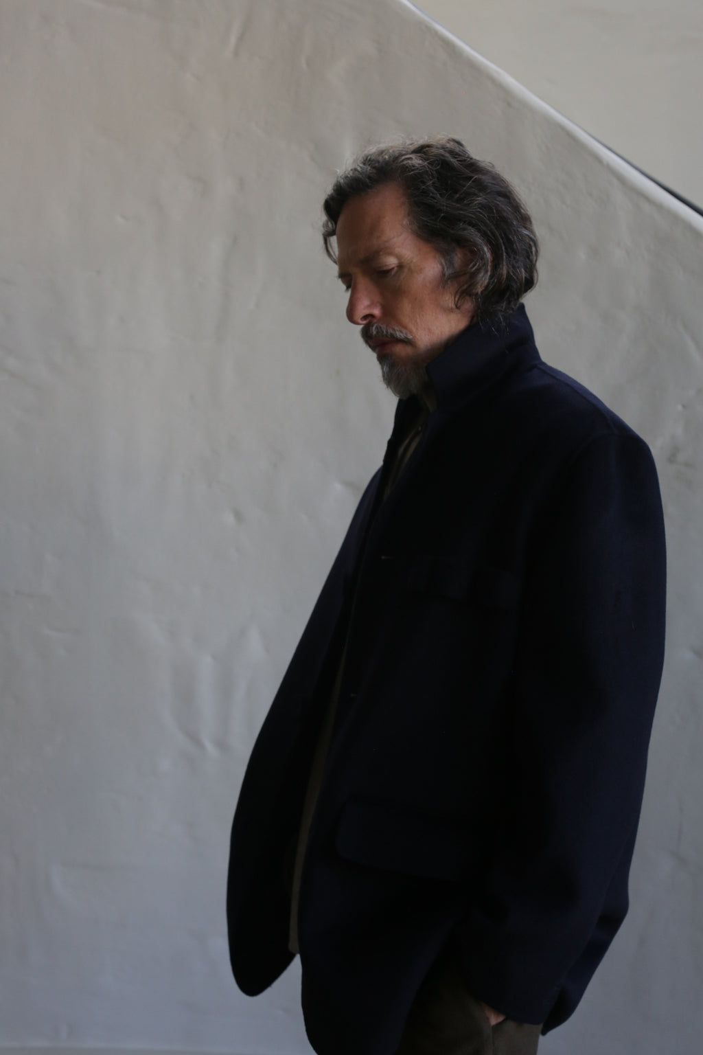 A man with wavy hair and a beard stands against a light background, looking down thoughtfully. He wears the Sculptor's Jacket in navy double face cashmere—an elegant blend of practical design and refined style.