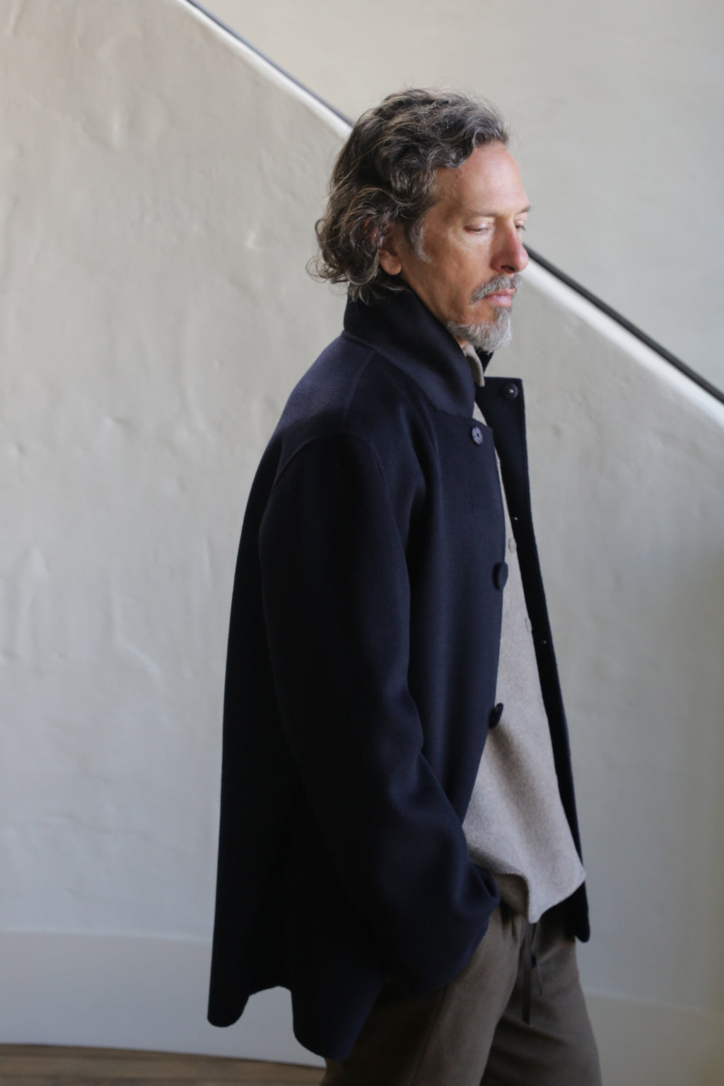 A man with curly hair and a goatee stands indoors against a light-colored textured wall, wearing the Sculptor's Jacket in Double Face Cashmere Navy over a beige sweater, looking down thoughtfully.