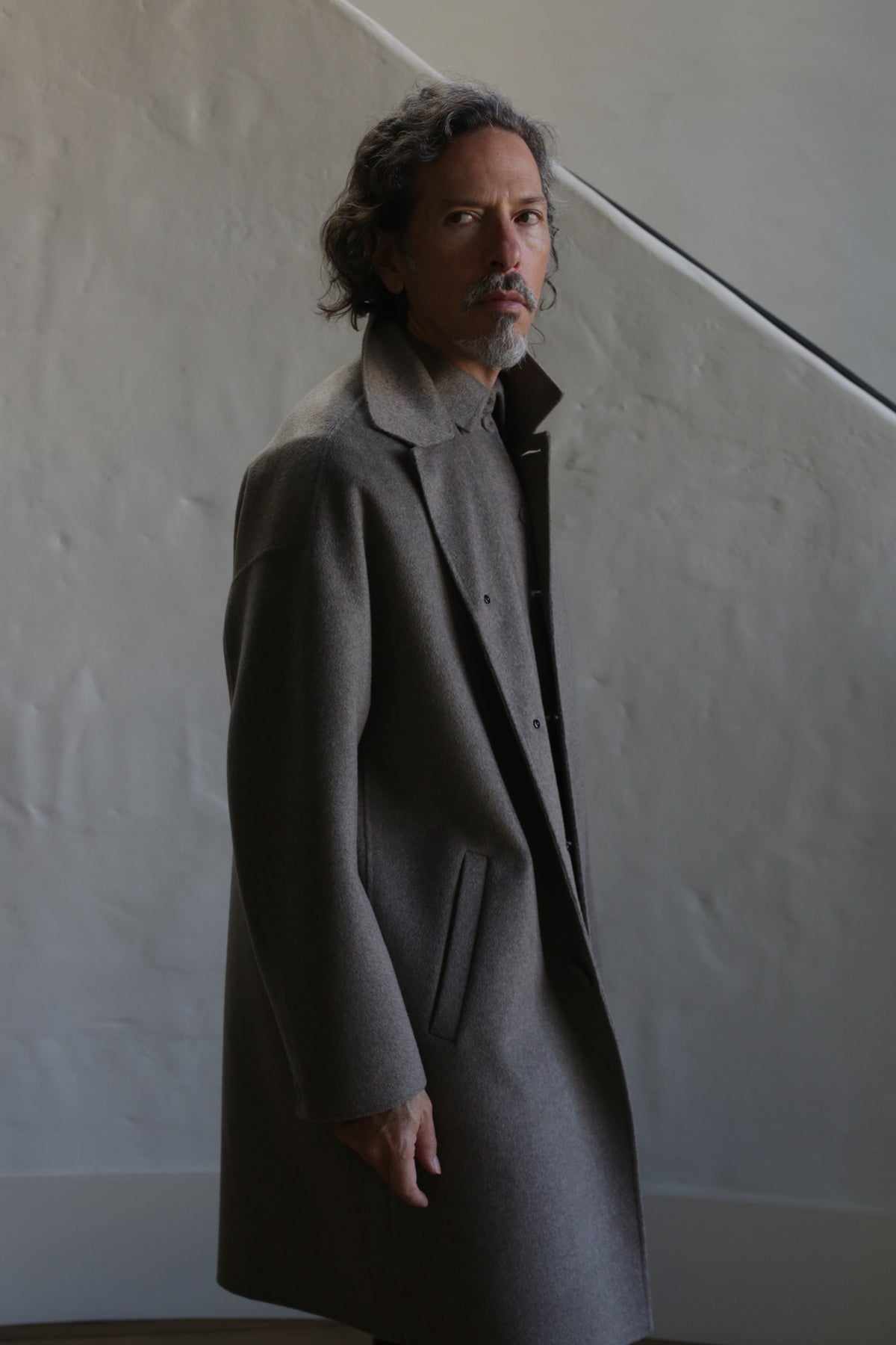 A man with wavy hair and a mustache wears the Men's Coat | Double Face Cashmere Undyed Natural indoors, standing against a textured, light-colored wall with a diagonal line in the background.