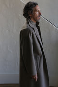A man with wavy dark hair and a short beard wears the Men's Coat | Double Face Cashmere Undyed Natural, standing indoors in profile against a textured light wall and staircase.