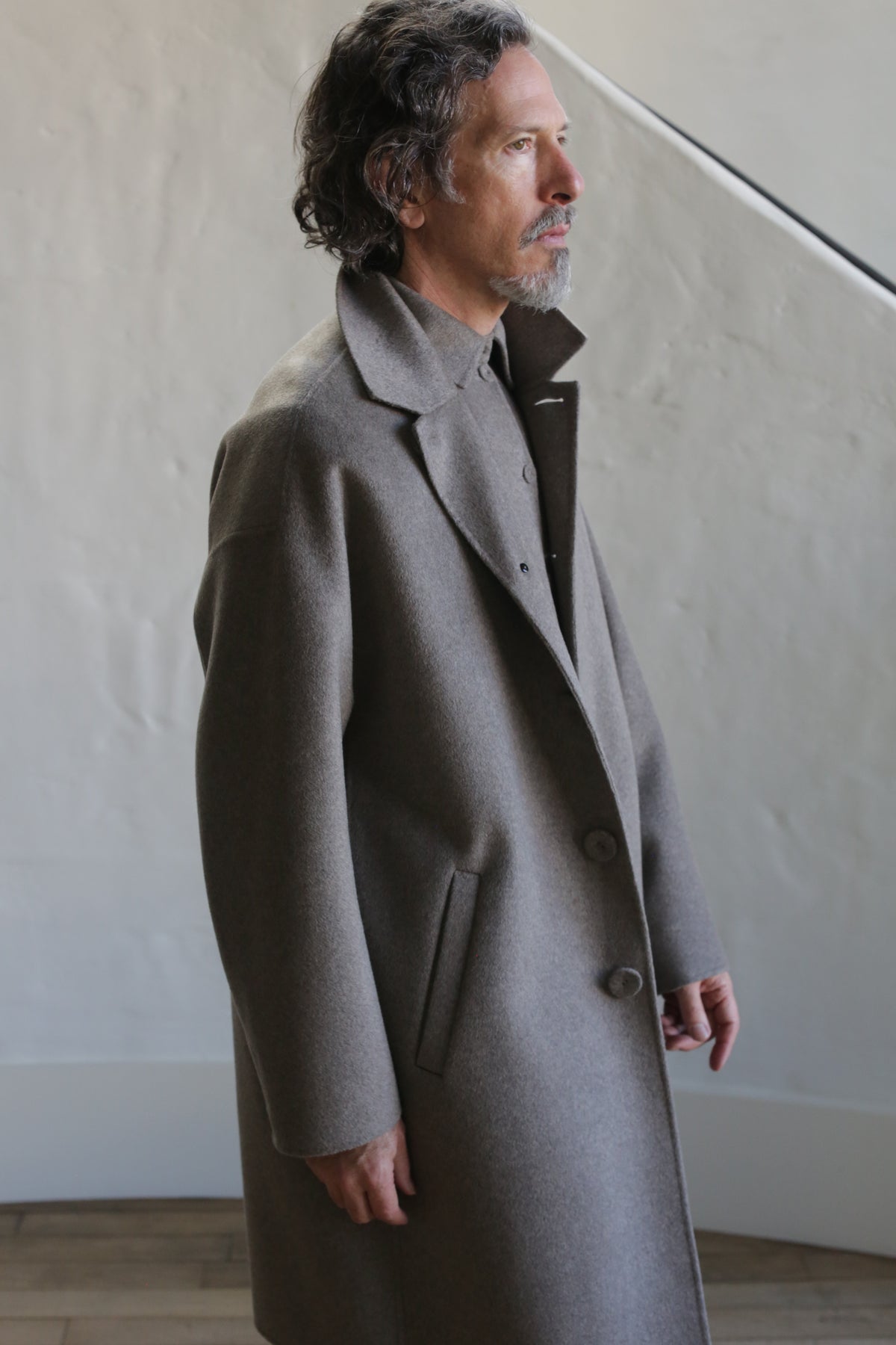A man with wavy hair and a short beard stands indoors wearing the Men's Coat | Double Face Cashmere Undyed Natural over a collared shirt, gazing to the side near a textured light wall and wooden floor.