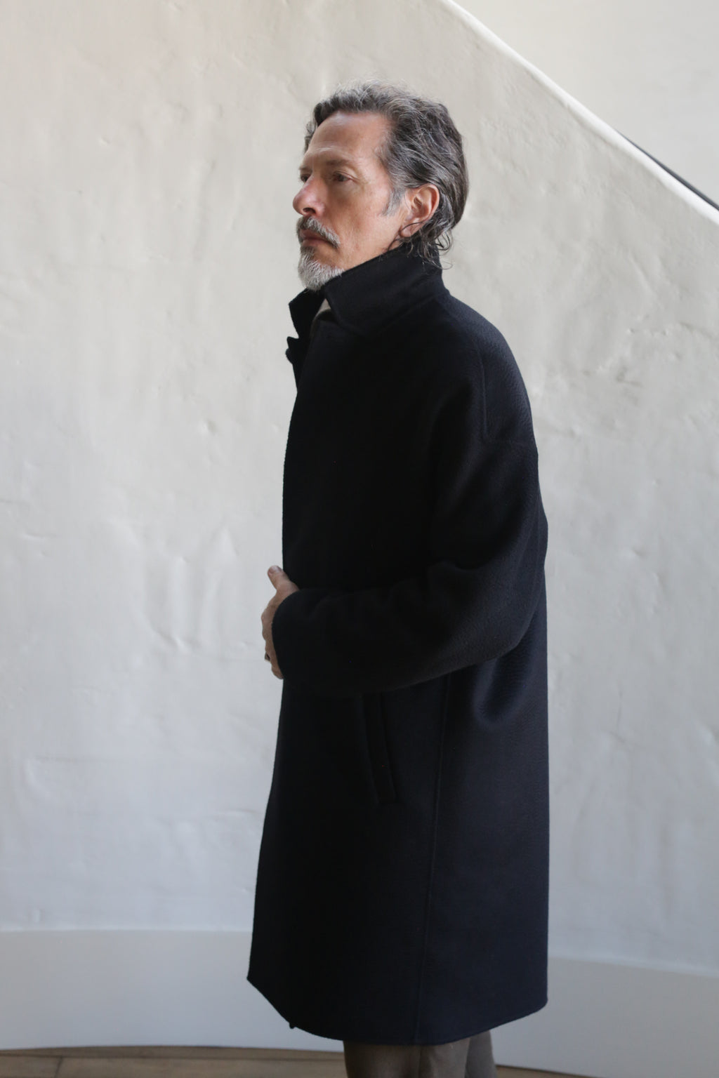 A gray-haired man with a mustache stands in profile against a white textured wall, wearing the Men's Coat | Watermark Double Face Cashmere Black with the collar turned up.