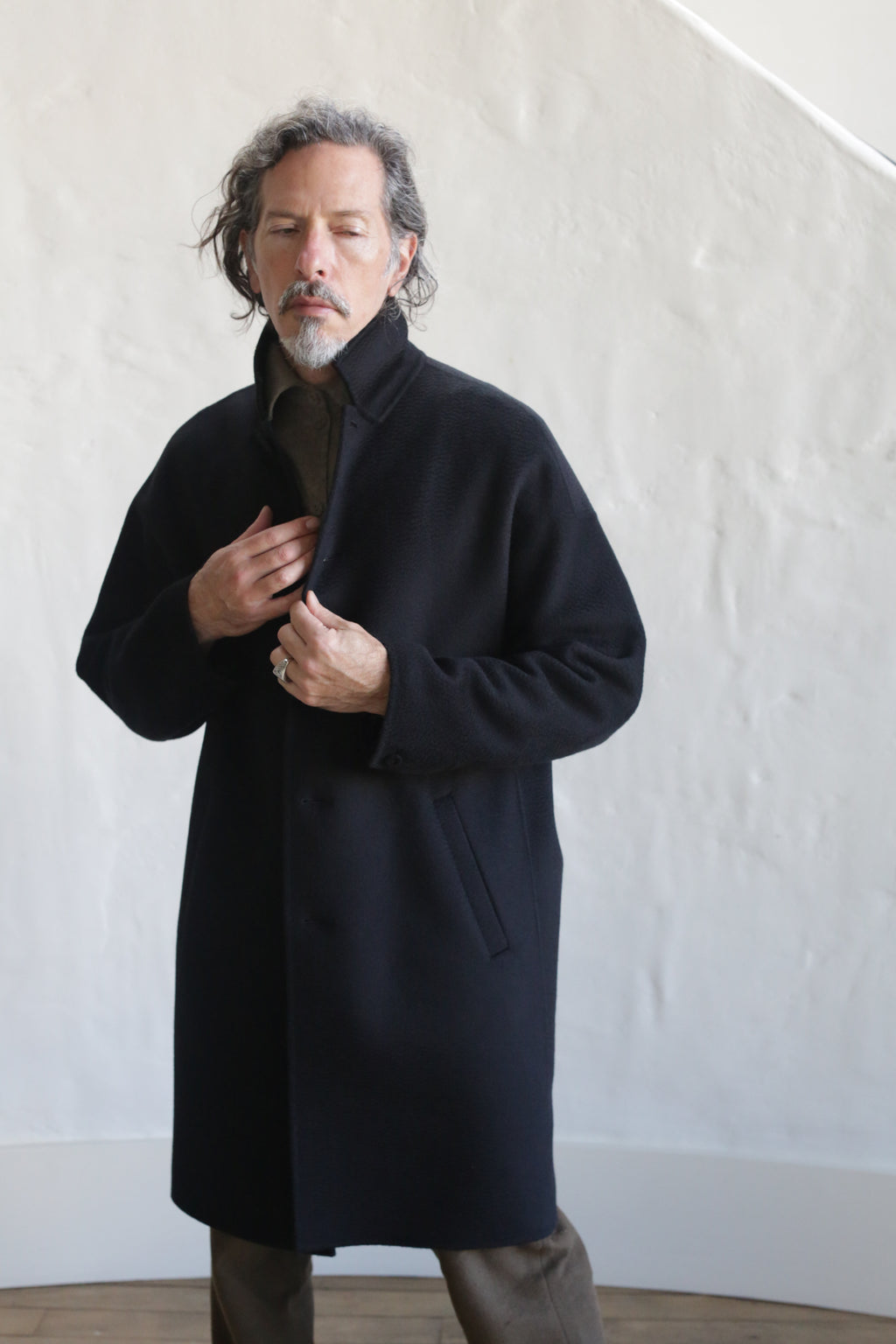 A man with wavy gray hair and a mustache stands indoors against a textured white wall, buttoning up the Men's Coat | Watermark Double Face Cashmere Black, looking down with a calm expression.