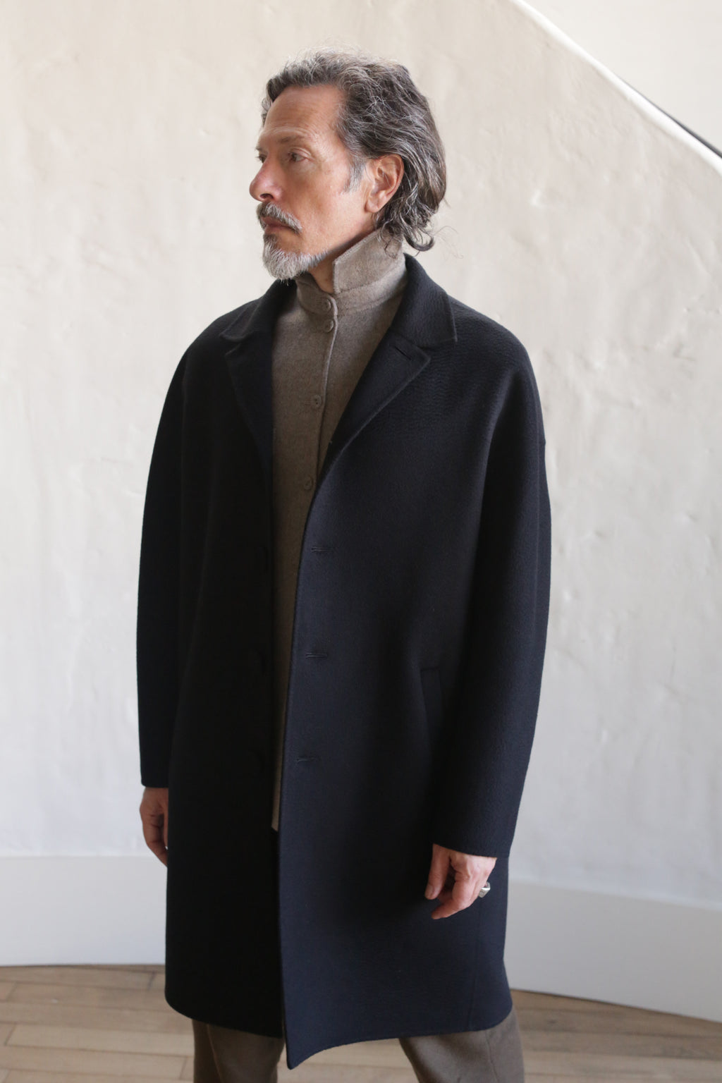 A man with gray hair and a mustache wears the Men's Coat | Watermark Double Face Cashmere Black over a light shirt, standing indoors against a textured white wall and wooden floor.