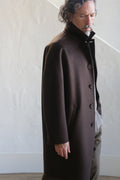 A man with wavy, graying hair and a mustache stands by a light wall, wearing the Men's Coat | Watermark Double Face Cashmere Brown over a gray shirt and olive pants. He looks down with a contemplative expression.