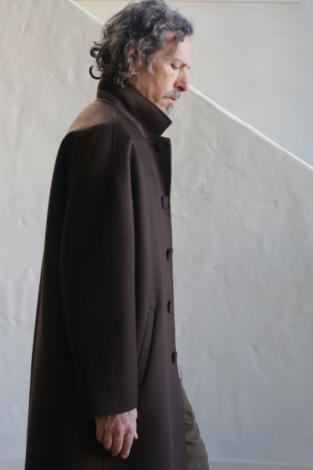 A man with wavy gray hair and a mustache wears the Men's Coat | Watermark Double Face Cashmere Brown, standing in profile against a light-colored textured wall with a diagonal shadow.