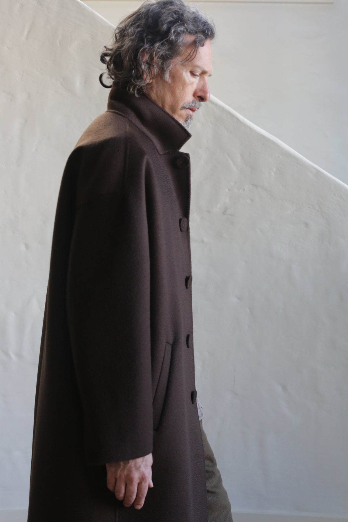 A man with wavy gray hair and a mustache wears the Men's Coat | Watermark Double Face Cashmere Brown, standing in profile against a light-colored textured wall with a diagonal shadow.