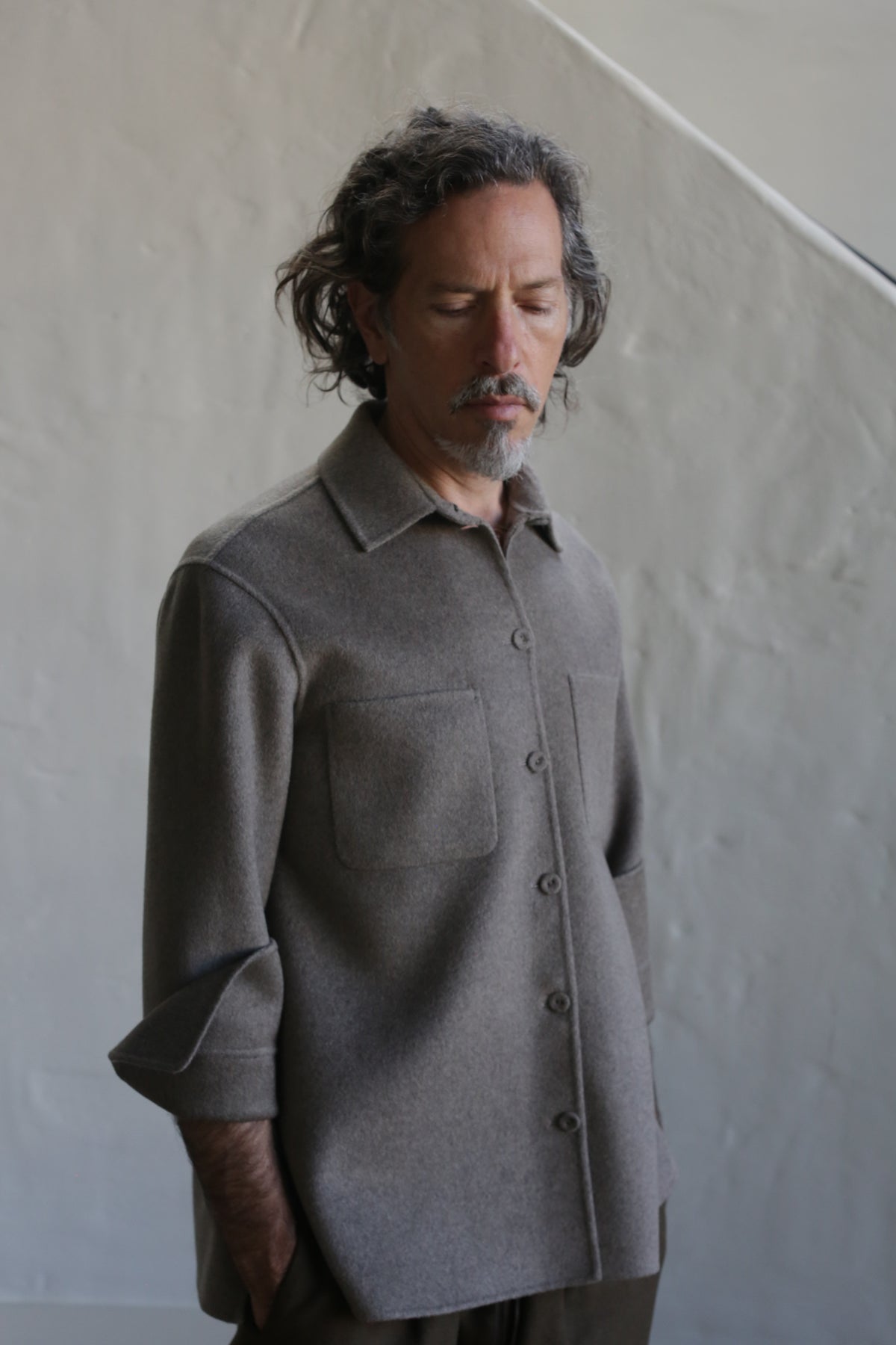 A man with wavy hair and a beard stands against a textured light-gray wall, wearing the Unisex Studio Shirt in undyed natural double face cashmere, his hands behind his back and eyes closed.