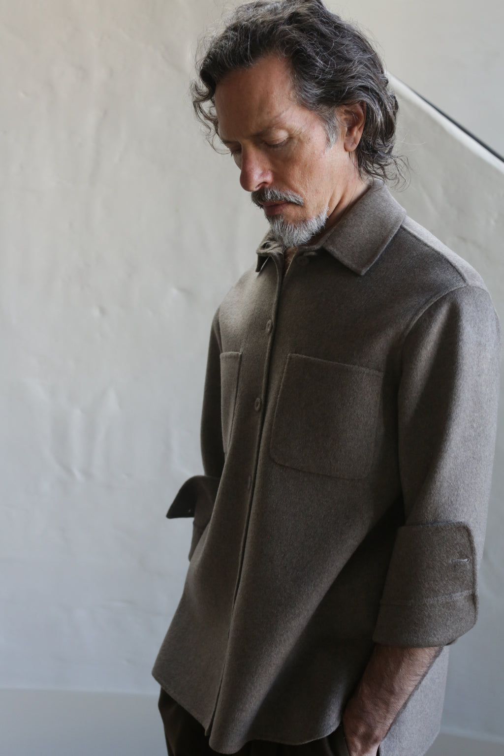 A man with wavy dark hair and a gray beard stands indoors, looking downward, wearing the Unisex Studio Shirt in double face undyed natural cashmere, featuring large chest pockets against a plain neutral background.