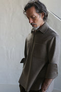 A man with wavy dark hair and a gray beard stands indoors, looking downward, wearing the Unisex Studio Shirt in double face undyed natural cashmere, featuring large chest pockets against a plain neutral background.