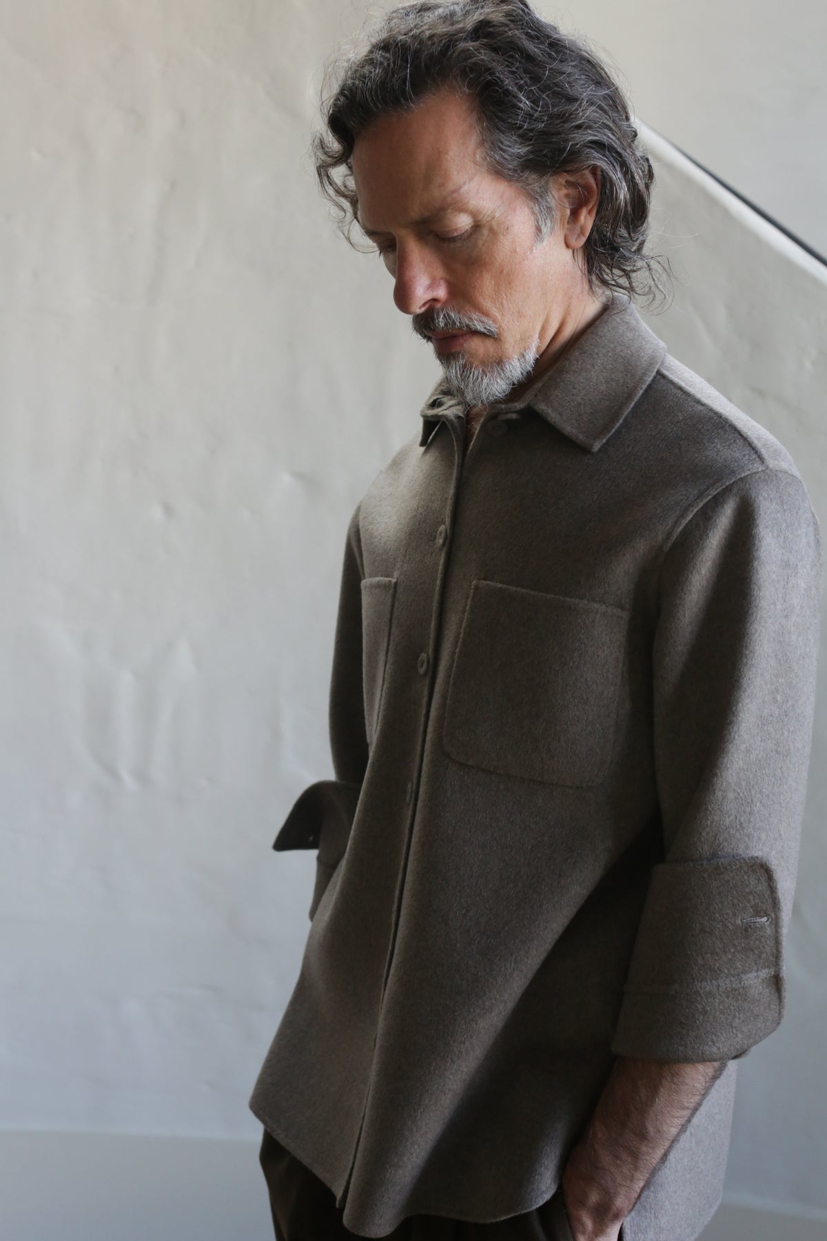 A man with wavy dark hair and a gray beard stands indoors, looking downward, wearing the Unisex Studio Shirt in double face undyed natural cashmere, featuring large chest pockets against a plain neutral background.