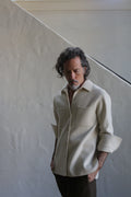 A man with wavy hair and a beard wears the Unisex Studio Shirt | Double Face Cashmere Undyed, featuring large pockets, paired with dark pants, standing hands-in-pockets before a light textured wall and diagonal staircase.