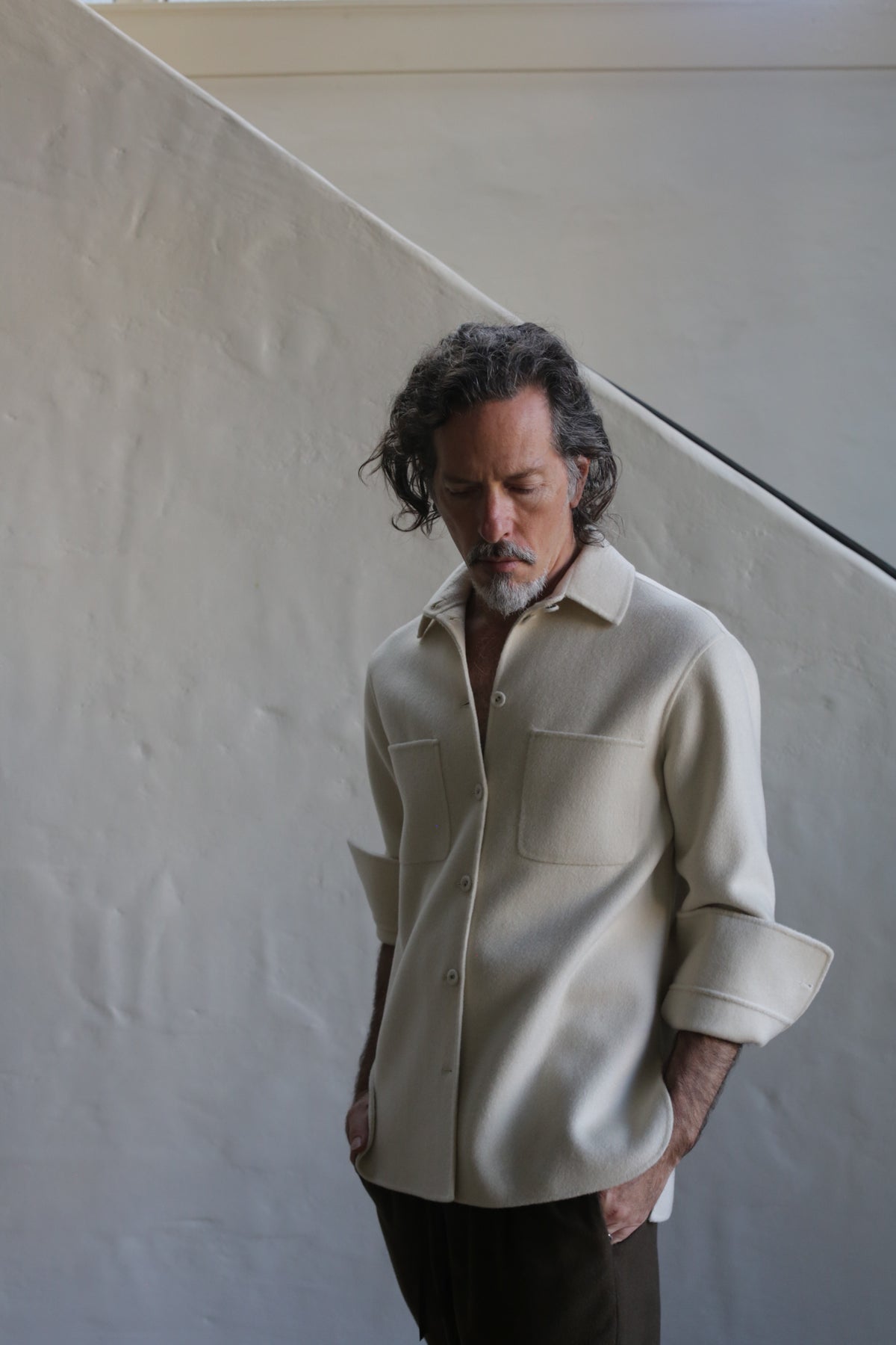 A man with wavy hair and a beard wears the Unisex Studio Shirt | Double Face Cashmere Undyed, featuring large pockets, paired with dark pants, standing hands-in-pockets before a light textured wall and diagonal staircase.