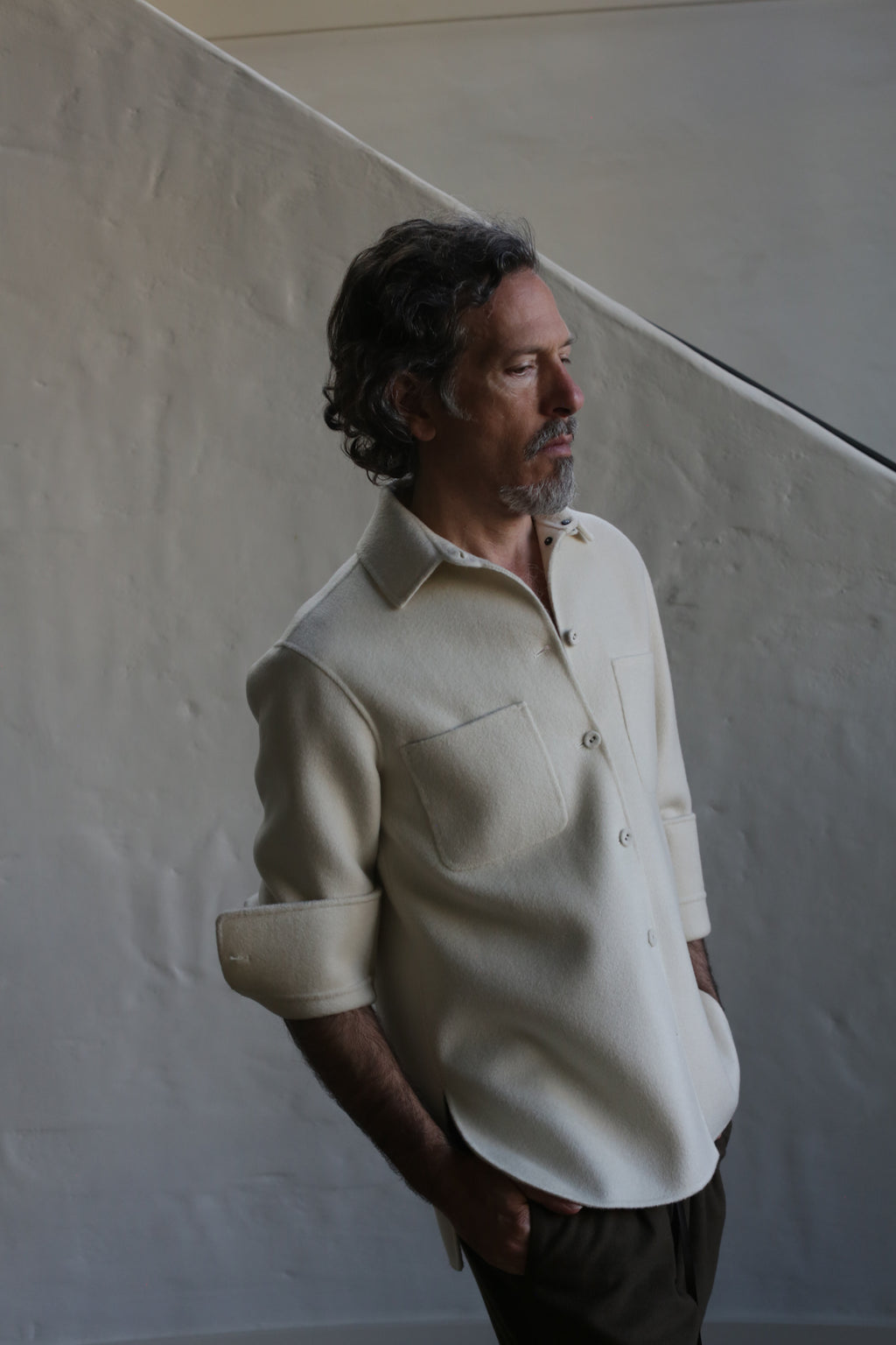 A man with wavy gray hair and a beard stands sideways by a light-colored wall and staircase, hands in pockets. He wears the Unisex Studio Shirt | Double Face Cashmere Undyed with rolled-up sleeves, looking thoughtful.