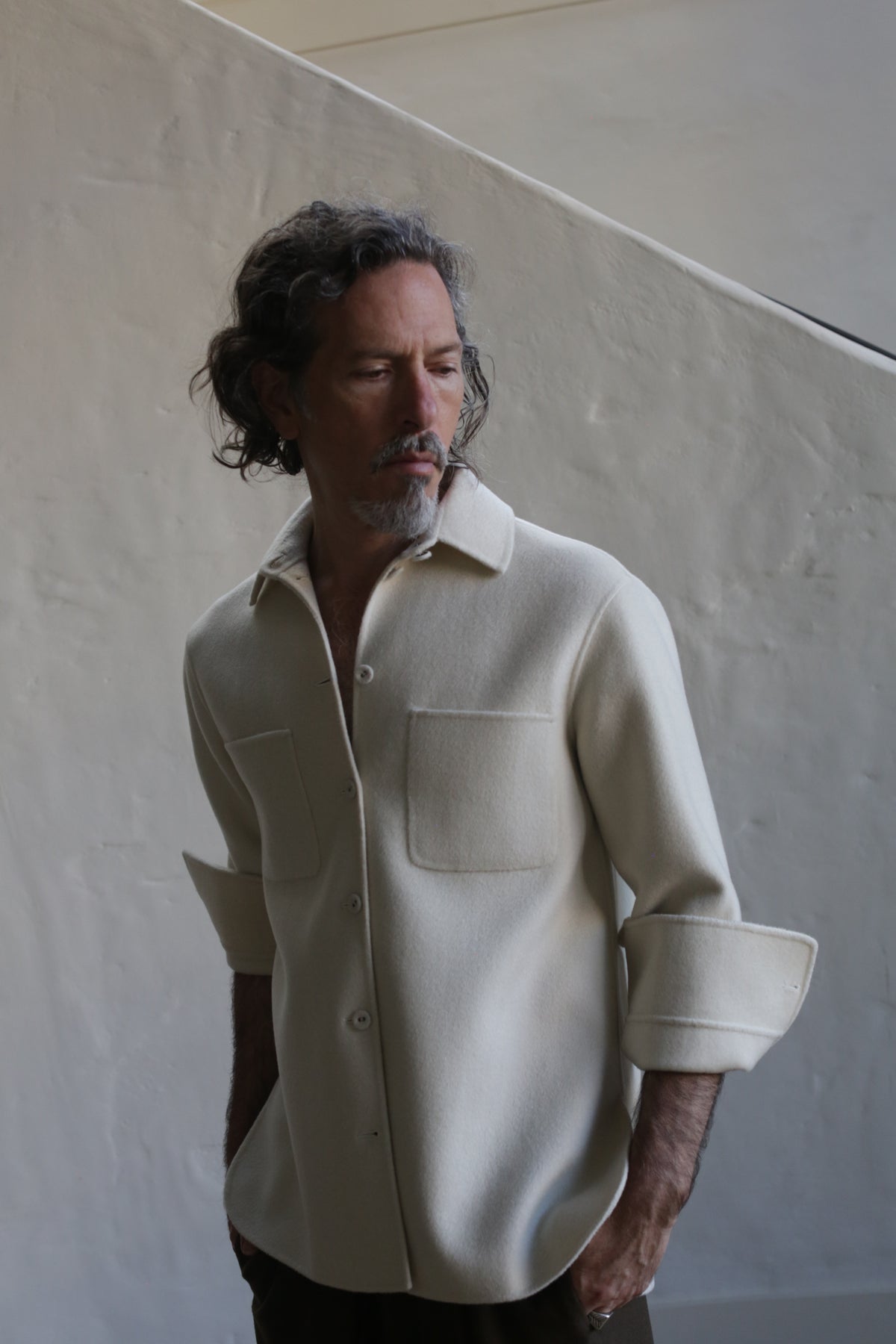 A man with wavy gray hair and a beard wears the Unisex Studio Shirt in Double Face Cashmere Undyed, featuring large front pockets. He stands before an off-white wall, looking down to his side with a thoughtful expression.