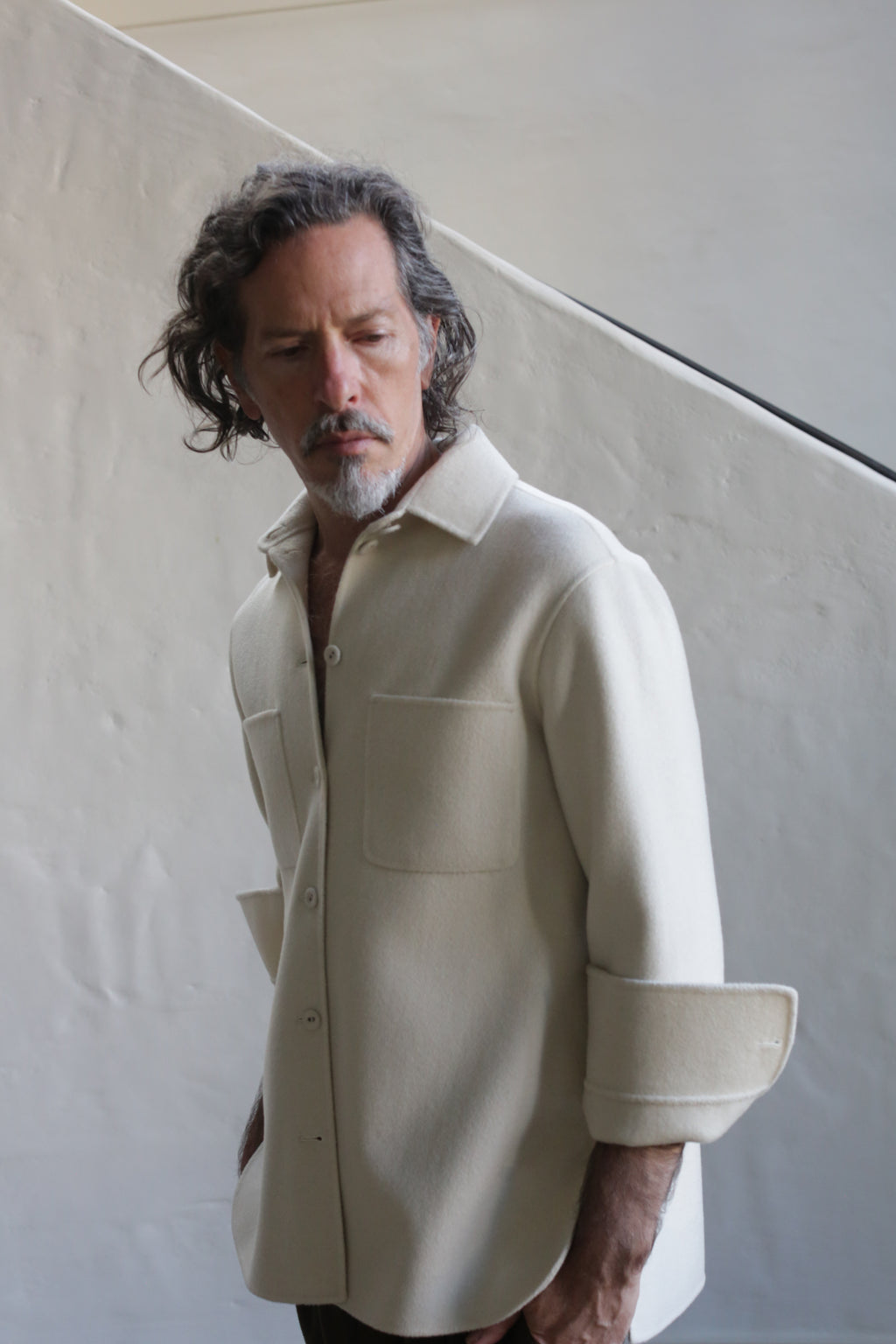A man with wavy, shoulder-length hair and a mustache wears the Unisex Studio Shirt in Double Face Cashmere Undyed, sleeves rolled up, standing before a textured light wall and looking downward.