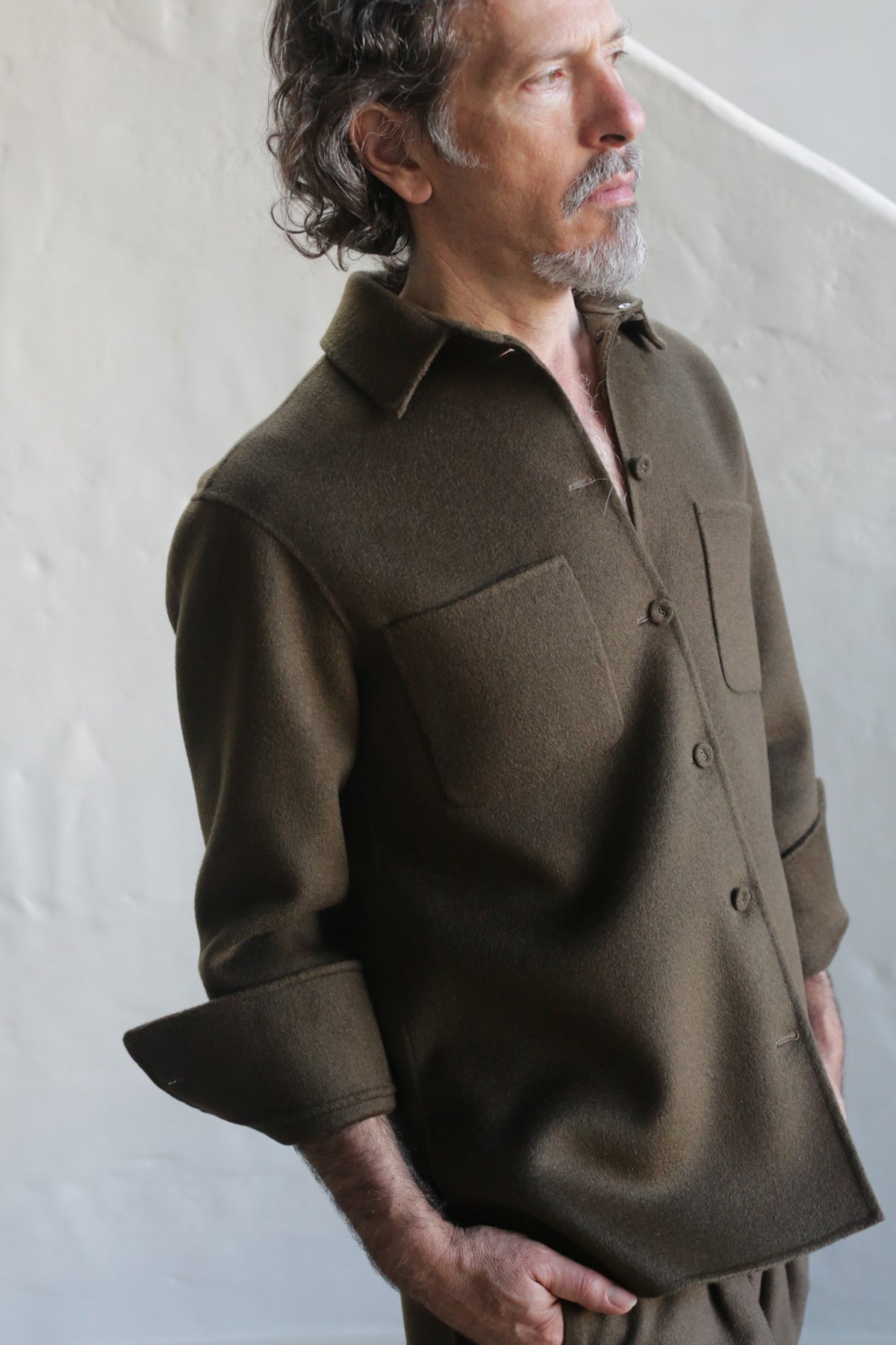 A man with wavy hair and a short beard looks to the side, wearing the Unisex Studio Shirt | Double Face Cashmere Olive, featuring large chest pockets and rolled-up sleeves, against a light-colored textured wall.