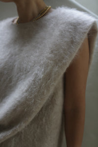 Close-up of a person wearing the Salento | Sandstone, a soft and fuzzy sleeveless top made from brushed cashmere in a light beige hue. The texture of the fabric is prominent, showcasing its fluffy and cozy appearance. The person also sports a gold necklace, partially visible against this luxurious vest. The background is blurred.