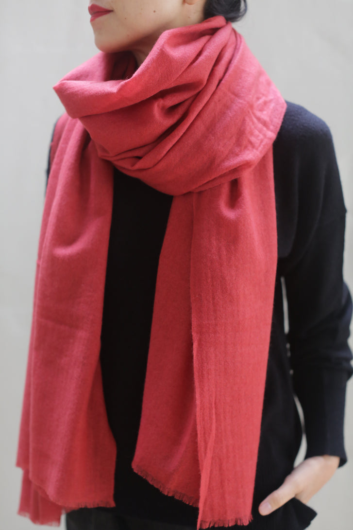 A person wearing a black long-sleeve top is shown from the shoulders down. They have wrapped a luxurious Tissue Weight Cashmere Scarf in Coral around their neck, with the ends draping down their front. The background is plain and light-colored.