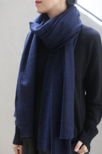 A person is wearing a Tissue Weight Cashmere Scarf in navy, draped around their neck over a black sweater. The person has their head turned slightly to the side, with only their lower face visible, showing red lipstick. The background is neutral and out of focus.