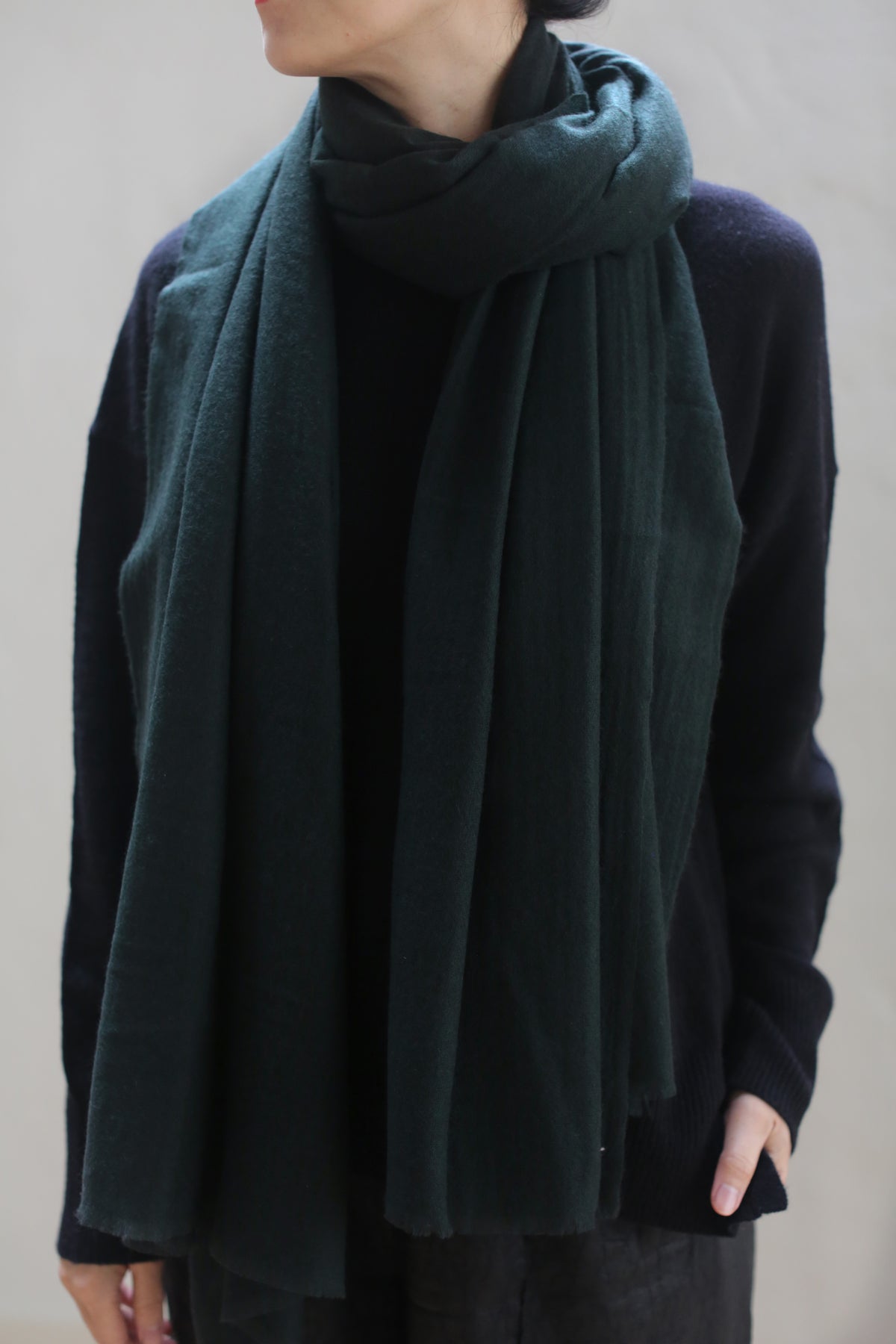 Person wearing a dark-colored sweater and a Hunter Green Tissue Weight Cashmere Scarf wrapped loosely around their neck. The image focuses on the upper body and neck, with the face partially hidden. The 36" x 82" cashmere scarf adds an elegant touch to the ensemble.
