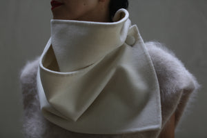 A person is wearing the stylish Wrap Scarf in off-white, made of undyed double-face cashmere, with a high collar that is artistically folded and accented with a button detail. Layered over it is a soft, fuzzy grey top. The background is a muted, solid color.