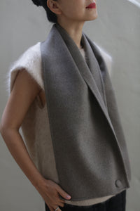 A person wearing a sleeveless beige top paired with the handmade Wrap Scarf in Undyed Natural from Atelier Lan Janicke draped around their neck. The person's head is slightly turned to the side, and only part of their face is visible. They have short dark hair, and the background is plain and muted.