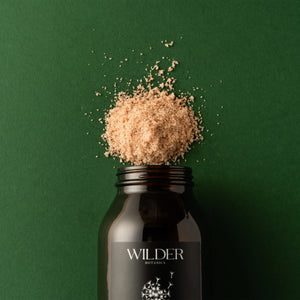 A dark glass jar of Healing Spirit Soak salt scrub, enriched with Pink Himalayan salts, is set against a solid green background. The jar's black label features white text and a floral design. The scrub is heaped on top of the open jar, spilling slightly onto the background.