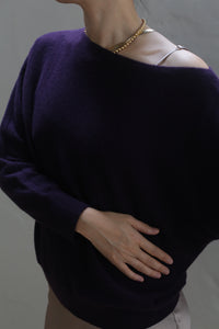 A person wearing the Ballerina's Cashmere Sweater in Aubergine, a true wardrobe staple, and a gold necklace is shown from the neck down, posing with one hand on their stomach against a plain background.