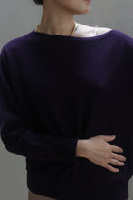 Load image into Gallery viewer, A person wearing the Ballerina&#39;s Cashmere Sweater in Aubergine with a boat neckline and a gold chain necklace stands with one arm resting across their stomach. Their face is turned slightly to the side, partially visible.