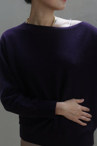 A person wearing the Ballerina's Cashmere Sweater in Aubergine with a boat neckline and a gold chain necklace stands with one arm resting across their stomach. Their face is turned slightly to the side, partially visible.