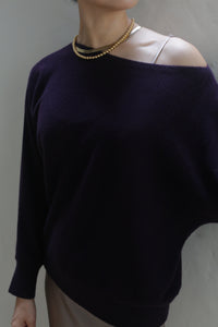 A person is wearing a Ballerina's Cashmere Sweater in aubergine, revealing a beige strap underneath, paired with a gold bead necklace. The background is a neutral gray wall, highlighting this wardrobe staple.