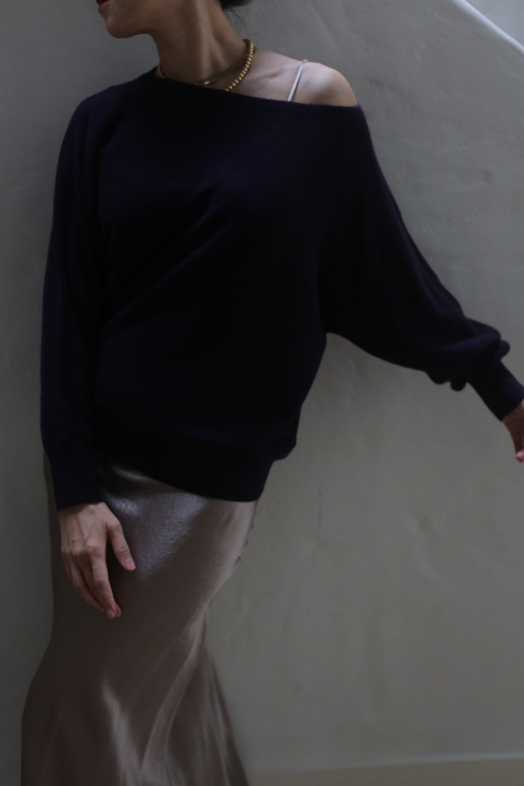 Clad in the Ballerina's Cashmere Sweater in Aubergine and a shimmering skirt, an individual poses against a plain wall. Their right arm is gently extended, and their head is turned, partially out of the frame. This sophisticated ensemble flawlessly merges comfort with elegance.